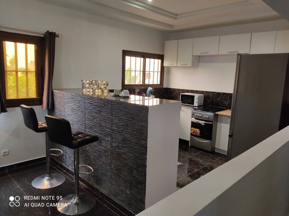 A modern kitchen space is visible, featuring a granite countertop with bar stools. Cabinets are arranged above and below the counter, and stainless steel appliances, including a refrigerator and microwave, are present. Natural light filters through the windows adorned with simple curtains.