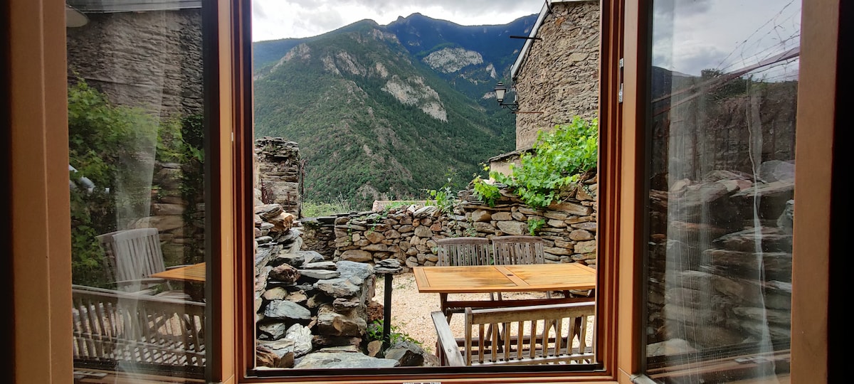 A view of the peaceful outdoor terrace is depicted, featuring a wooden table and benches beside a rustic stone wall. The backdrop showcases green mountains and a cloudy sky, inviting a connection with nature from inside the studio.