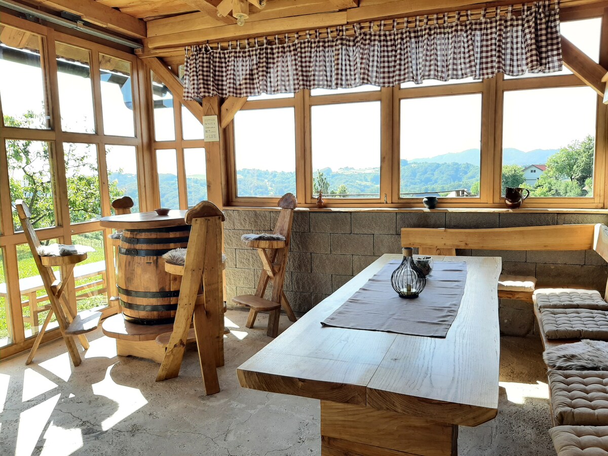 A sunlit dining area features large windows that frame scenic views of surrounding hills. The space includes a wooden table with cushioned benches, a barrel-shaped bar with stools, and decorative elements enhancing the natural ambiance.