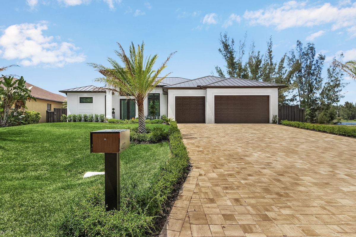 A modern single-story home with a sleek metal roof is highlighted against a bright blue sky. A spacious driveway made of pavers leads to a double garage. Lush green landscaping includes palm trees and manicured grass, creating an inviting entrance.