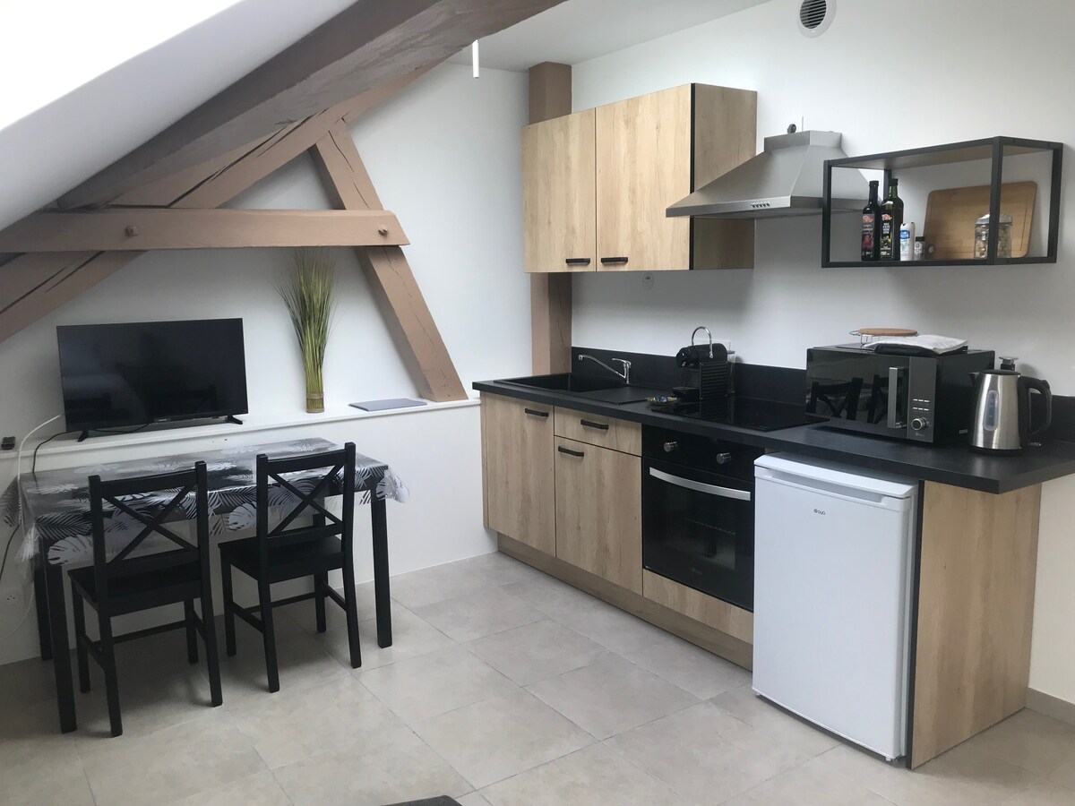 A modern kitchen area features light wood cabinets and a black countertop. A small dining table with four chairs is situated alongside a television. Appliances include a kettle, microwave, and refrigerator, with natural light illuminating the space through a nearby window.