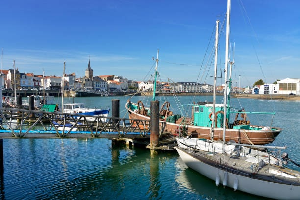 The image captures a serene harbor scene with several boats docked. A wooden boat with teal accents is prominently featured alongside a sleek white sailboat. Calm waters reflect the blue sky and buildings in the background, offering a glimpse of a quaint coastal town.