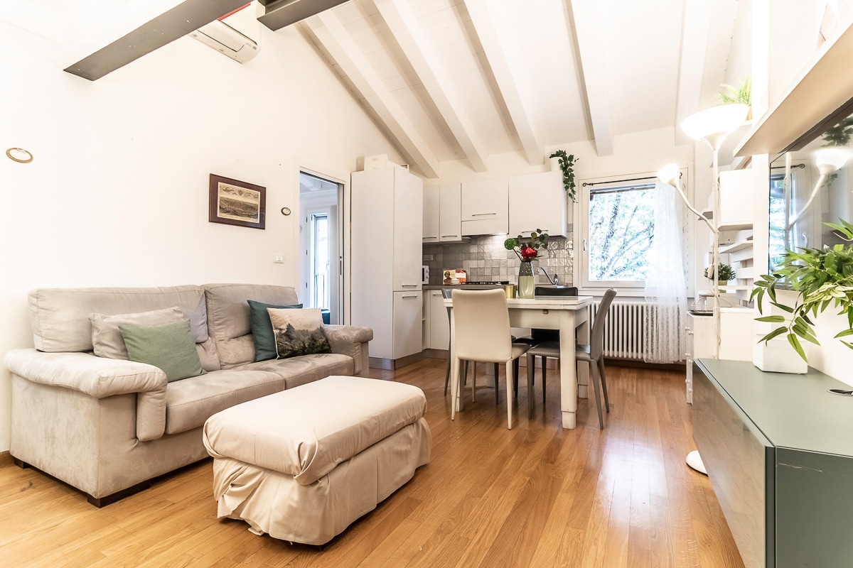 The living area includes a spacious gray sofa and a light-footed footrest, complemented by a dining table surrounded by four chairs. Soft natural light filters through a window adorned with sheer curtains, highlighting wooden flooring and minimalist accents, creating a comfortable gathering space.
