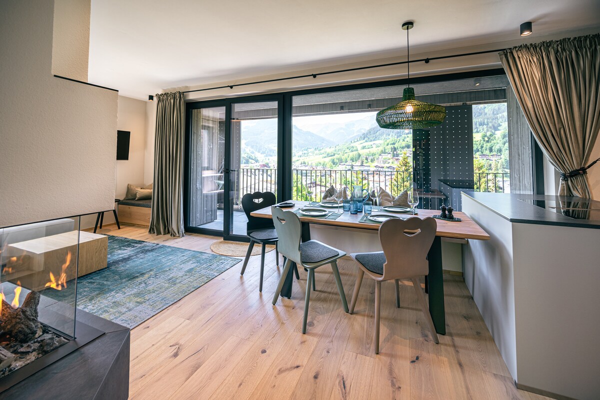 A bright dining area is furnished with a wooden table set for meals, surrounded by stylish chairs. Large glass doors open to a balcony with mountain views, while a cozy seating area is visible in the background. Natural light enhances the warm wood flooring.