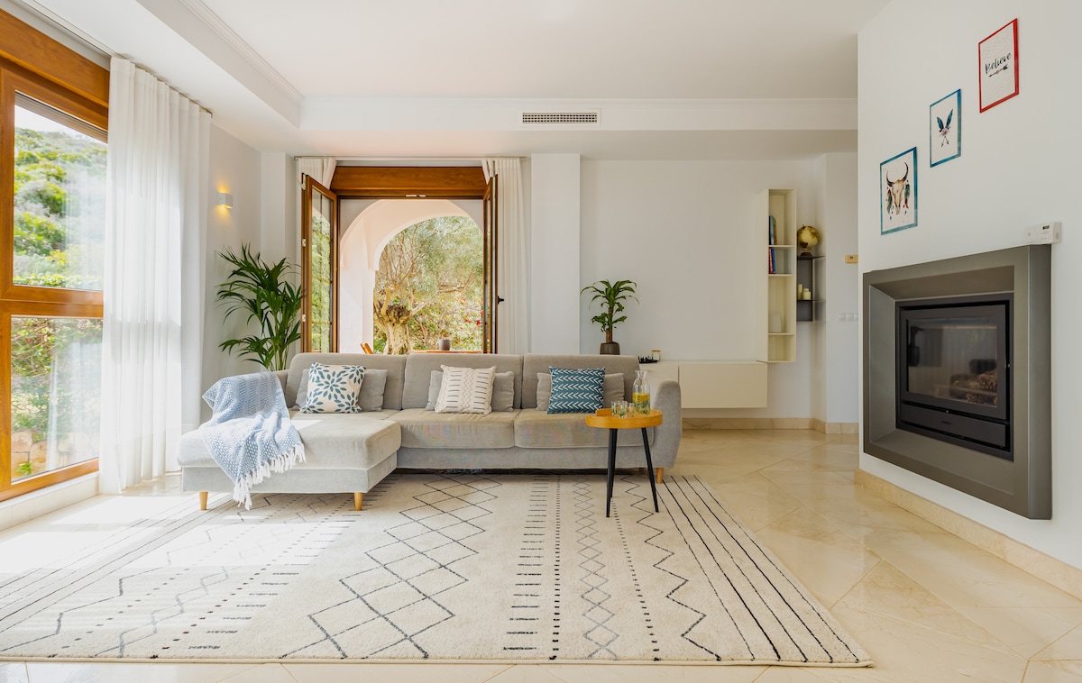A spacious living area features large windows that open to the outdoors, allowing natural light to fill the room. A comfortable sofa is accompanied by decorative pillows on a textured rug, and a simple coffee table rests nearby. A modern fireplace adds warmth to the ambiance.