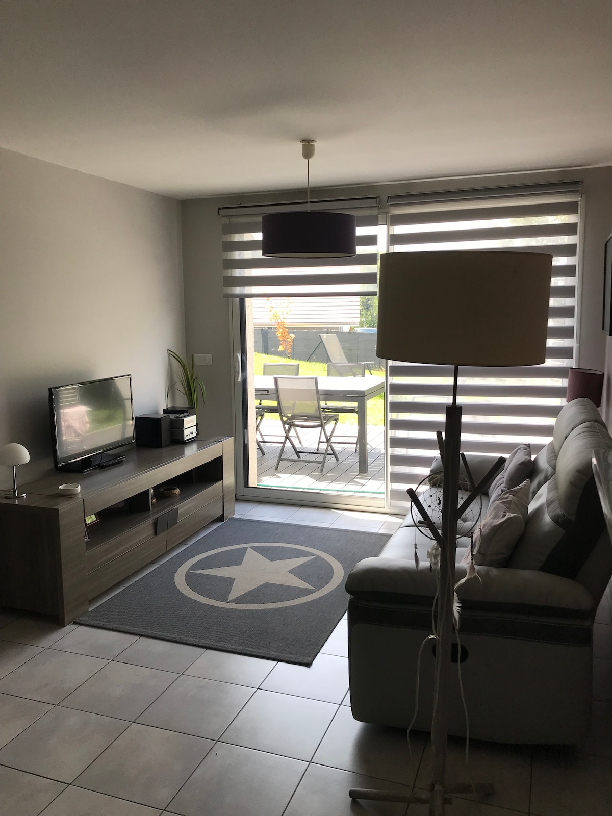A living area is displayed, featuring a comfortable sofa paired with a standing lamp. A television is positioned on a low cabinet. Large glass doors lead to a patio with outdoor seating, while light filters through dual roller shades.