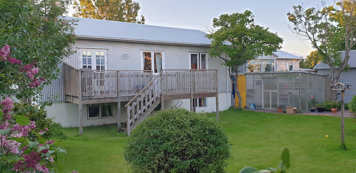 The exterior of a charming Scandinavian house is presented, showcasing a wooden deck that leads to the entrance. A well-maintained garden is visible, featuring green grass and leafy trees. Colorful flowers add vibrancy to the scene, while large windows reflect natural light.