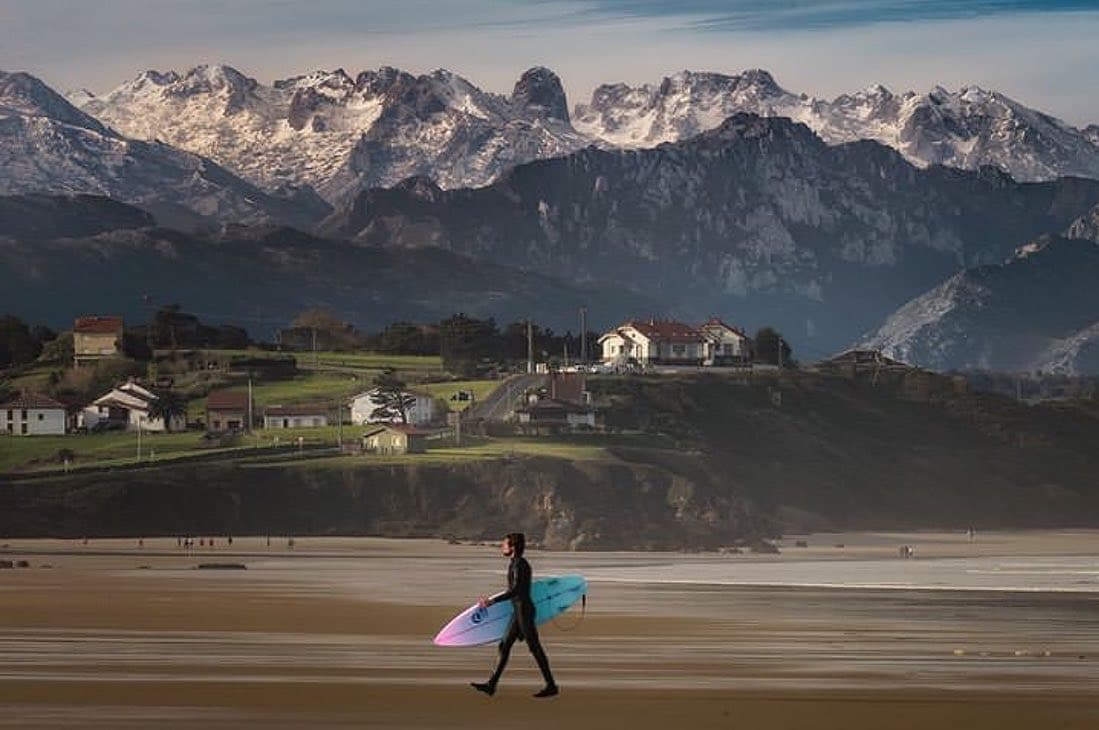 A surfer walks along the shoreline with a surfboard. Majestic snow-capped mountains rise in the background, framing a scenic coastal landscape. The beach is expansive with gentle waves lapping at the shore, creating a serene setting.