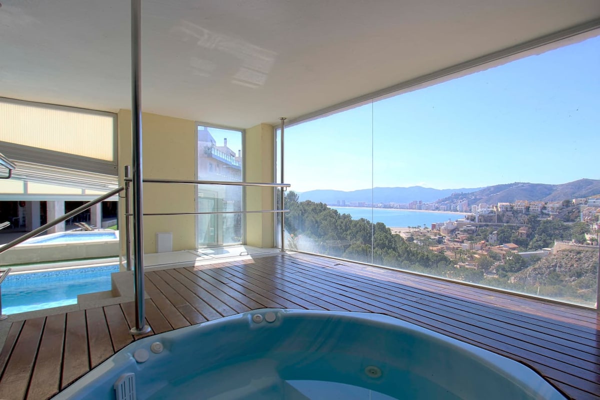 A spacious area features a jacuzzi with views of the bay through large glass windows. The wooden deck adds a natural element, creating a relaxing atmosphere with panoramic coastal scenery visible in the distance.