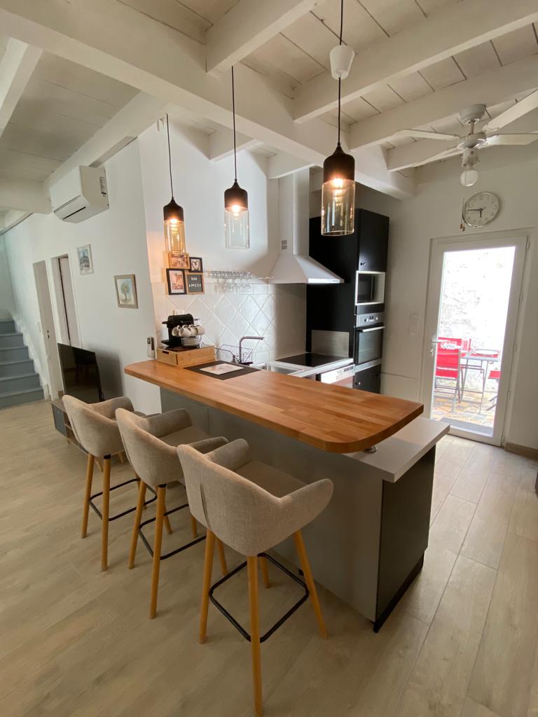 A modern kitchen area is highlighted by a wooden countertop, complemented by three upholstered bar stools. Stainless steel appliances and light-colored cabinets are visible. Natural light filters through a nearby door leading to an outdoor dining space.