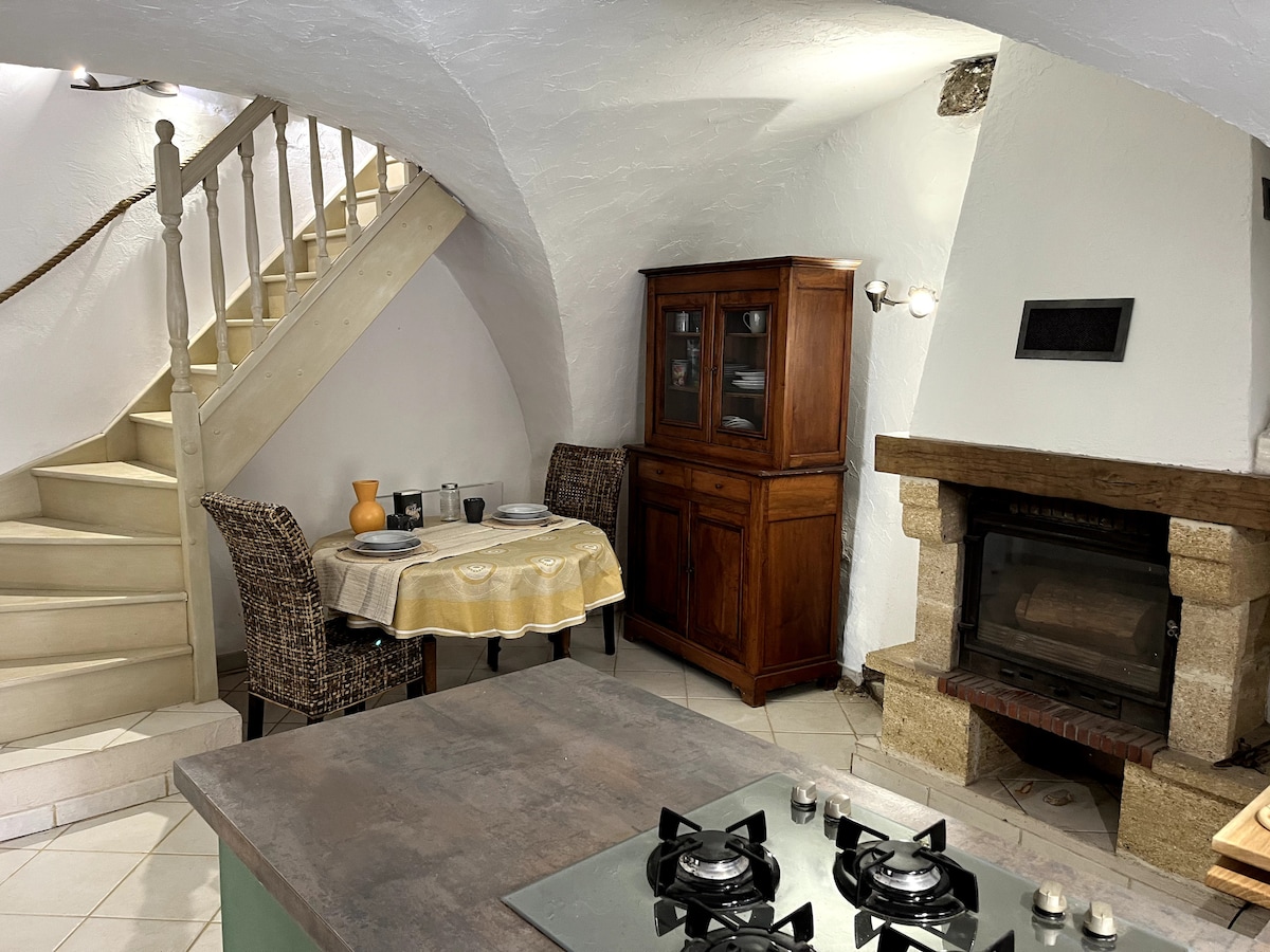 A cozy kitchen area features a wooden dining table set for two, accompanied by woven chairs. A stone fireplace is positioned adjacent to the dining space. In the background, a staircase leads to an upper level, while a sleek kitchen island with a stove completes the layout.