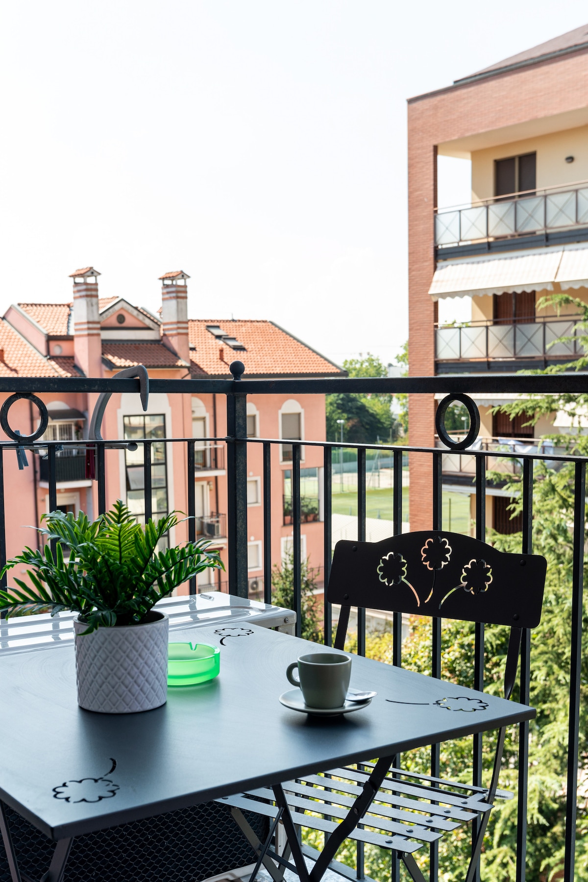 A balcony view features a metal table set with a small green plant and a cup. Potted greenery sits on the table, while the background includes a glimpse of neighboring buildings with balconies, all under a bright sky.