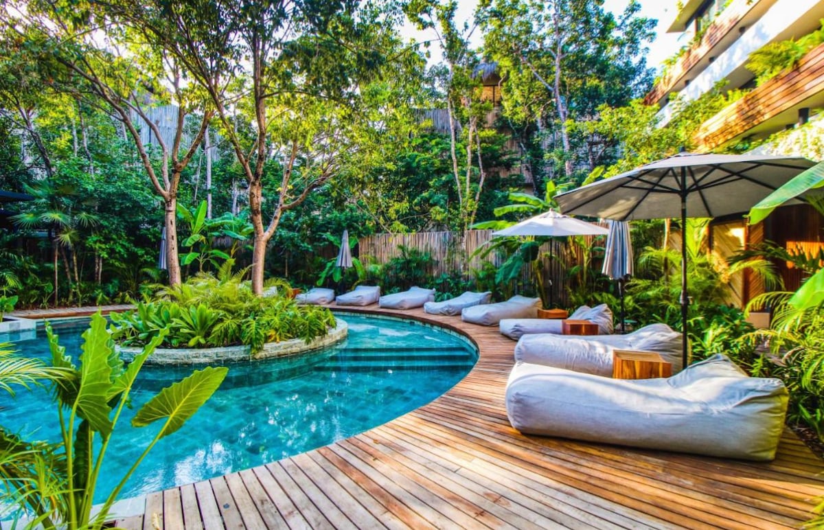 An inviting outdoor area presents a winding pool surrounded by lush greenery. Light-colored bean bag chairs are positioned on a wooden deck, with umbrellas offering shaded spots. The scene captures a tranquil atmosphere, enhanced by the natural elements of the surrounding foliage.