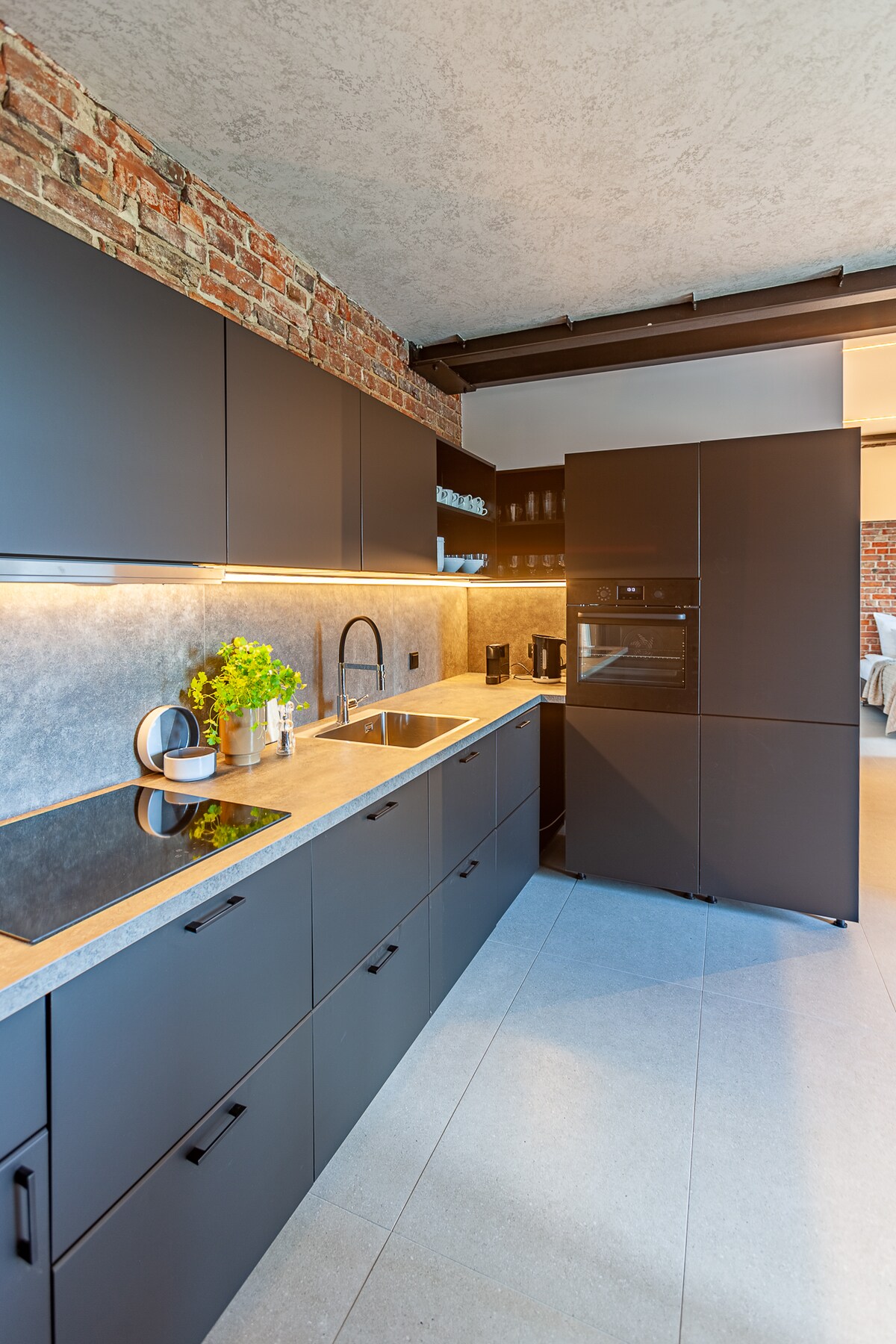 A modern kitchenette features sleek black cabinetry paired with a contrasting light countertop. A stainless steel sink and faucet are centrally placed. A built-in oven is situated next to a spacious refrigerator, with soft lighting highlighting the exposed brick wall.
