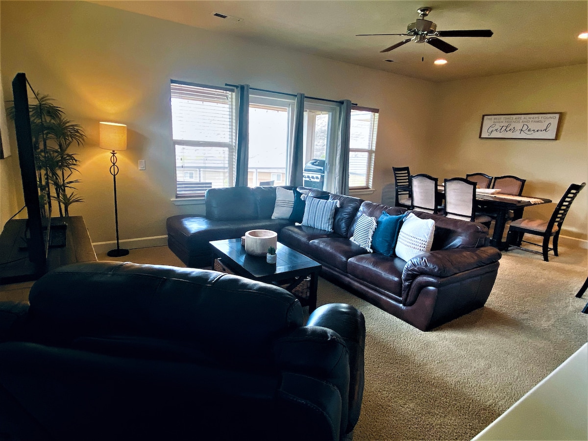 A spacious living area features a dark leather sectional sofa adorned with blue and white accent pillows. Natural light pours in through multiple windows, while a nearby dining table is visible. A lamp stands beside the sofa, contributing to the cozy atmosphere.