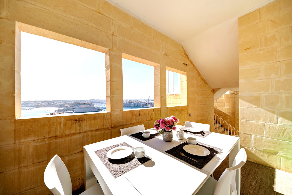 A bright dining area features a modern white table set for four, with elegant place settings and a vase of flowers at the center. Large windows framed in light-colored stone provide scenic views of the surrounding landscape, enhancing the open and airy atmosphere.