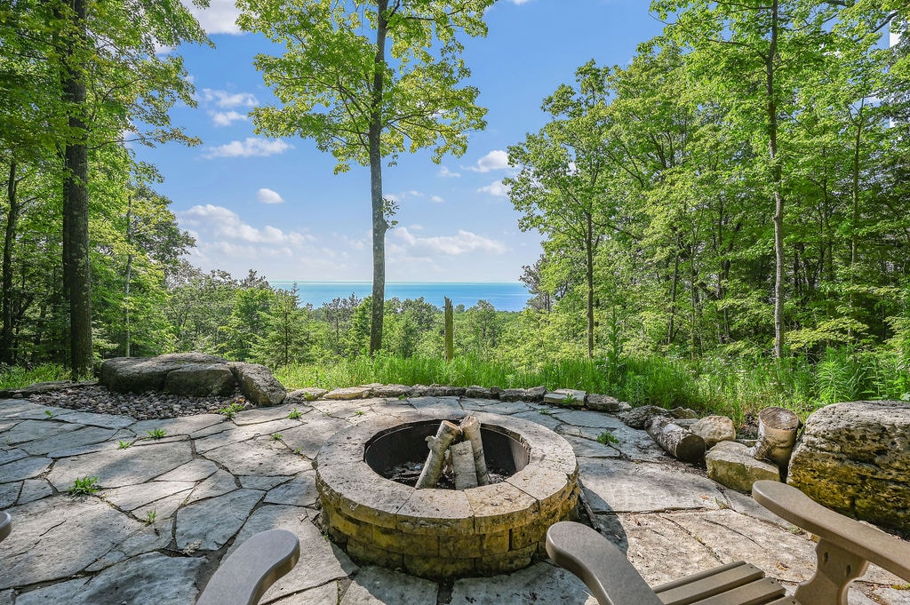 A stone fire pit is centered in the image, surrounded by natural stone seating and lush green foliage. Trees create a serene backdrop, with glimpses of the bay visible through the leaves, highlighting a tranquil outdoor setting perfect for relaxation.