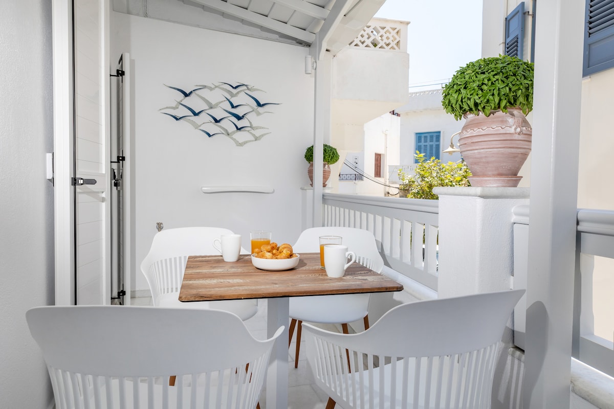 A balcony is presented with a wooden table and four white chairs. A bowl of pastries and two cups sit on the table. Decorative wall art featuring birds is displayed in the background, complemented by potted greenery and white architectural details.