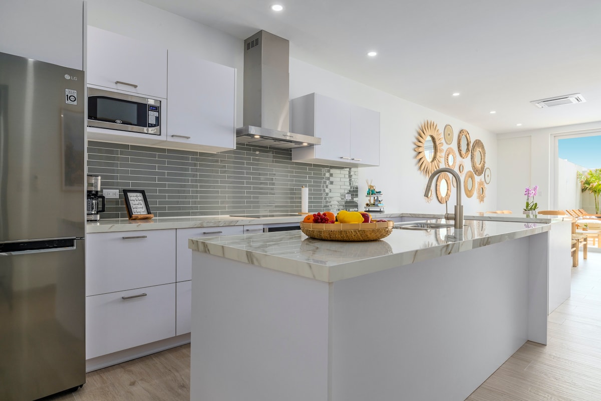 A modern kitchen displays sleek white cabinetry paired with a polished countertop. Stainless steel appliances complement the space, including a refrigerator, microwave, and oven. An island with a fruit bowl serves as a central feature, while natural light brightens the area through a nearby window.