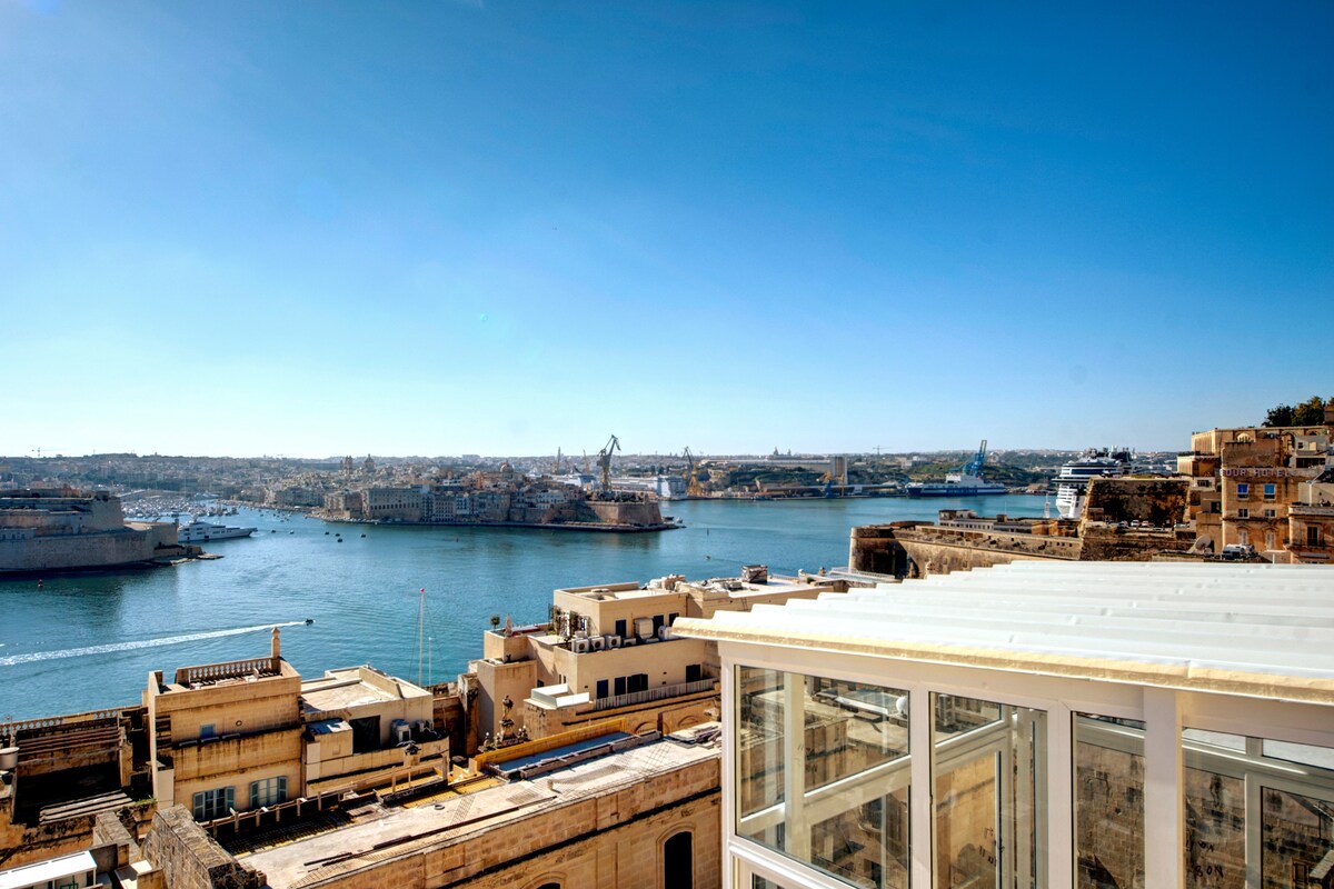 A breathtaking view of the grand harbour showcases the water glistening under the clear blue sky. Historical buildings and boats are visible in the distance, presenting a serene coastal scene. The foreground features modern architecture with glass, connecting the interior with the outdoor landscape.