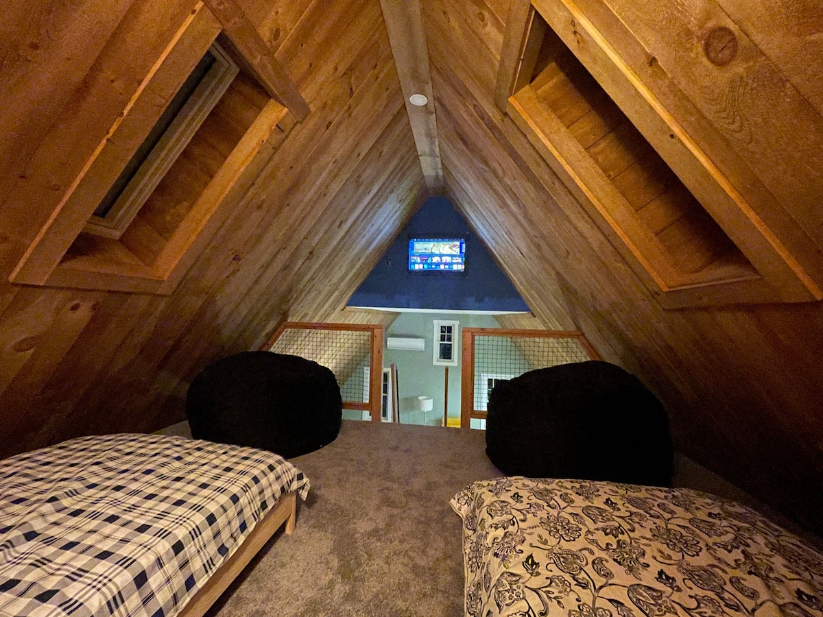 A cozy loft area is highlighted by slanted wooden ceilings and two openings allowing natural light. Two low beds with checkered and patterned bedding are positioned on a carpeted floor. A television is mounted on the far wall, completing the relaxing space.