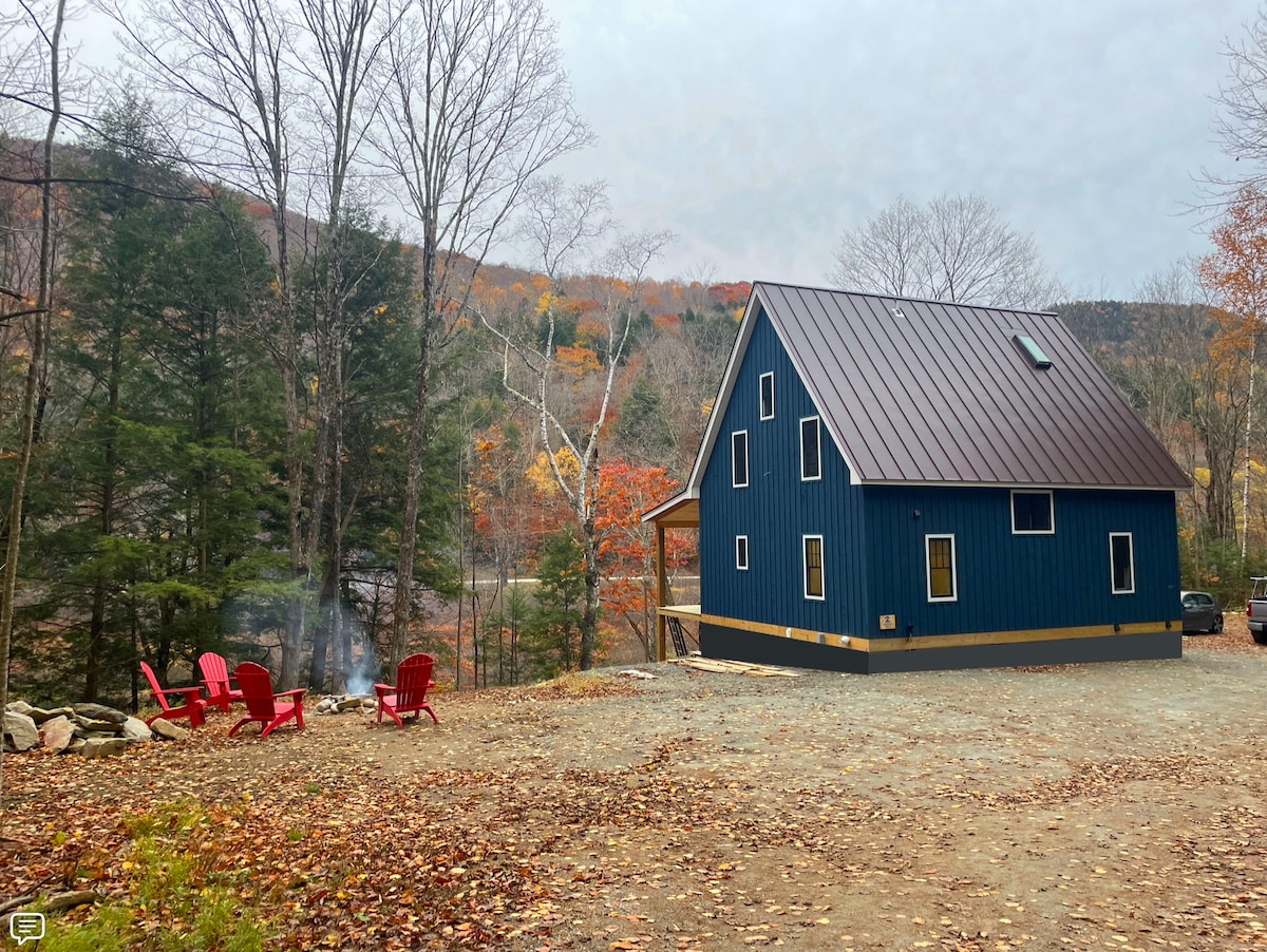 A modern two-story home features a dark blue exterior with a sloped metal roof. Comfortable red chairs surround a fire pit on a gravel area, surrounded by trees showcasing autumn colors. The backdrop includes a scenic view of the mountainous landscape.