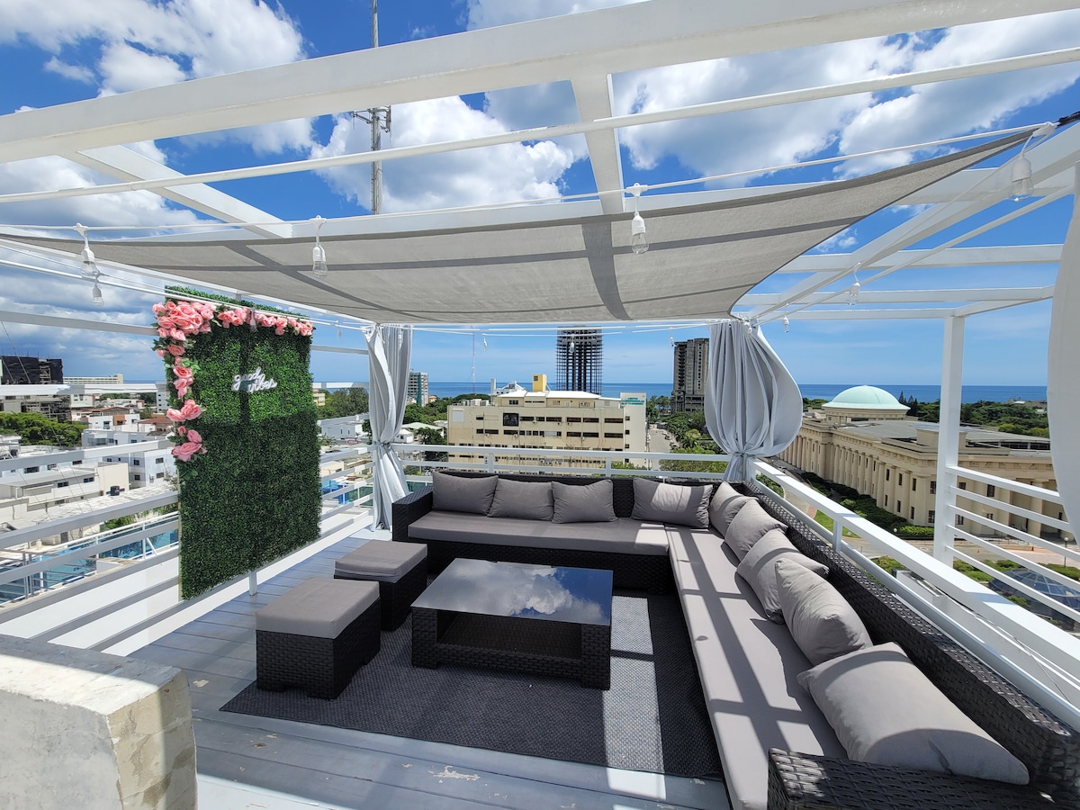 A spacious rooftop terrace features comfortable sectional seating, providing a relaxing area with stunning views of the ocean and city skyline. A decorative green wall adorned with flowers adds a natural touch, while an awning offers shade from the sun.
