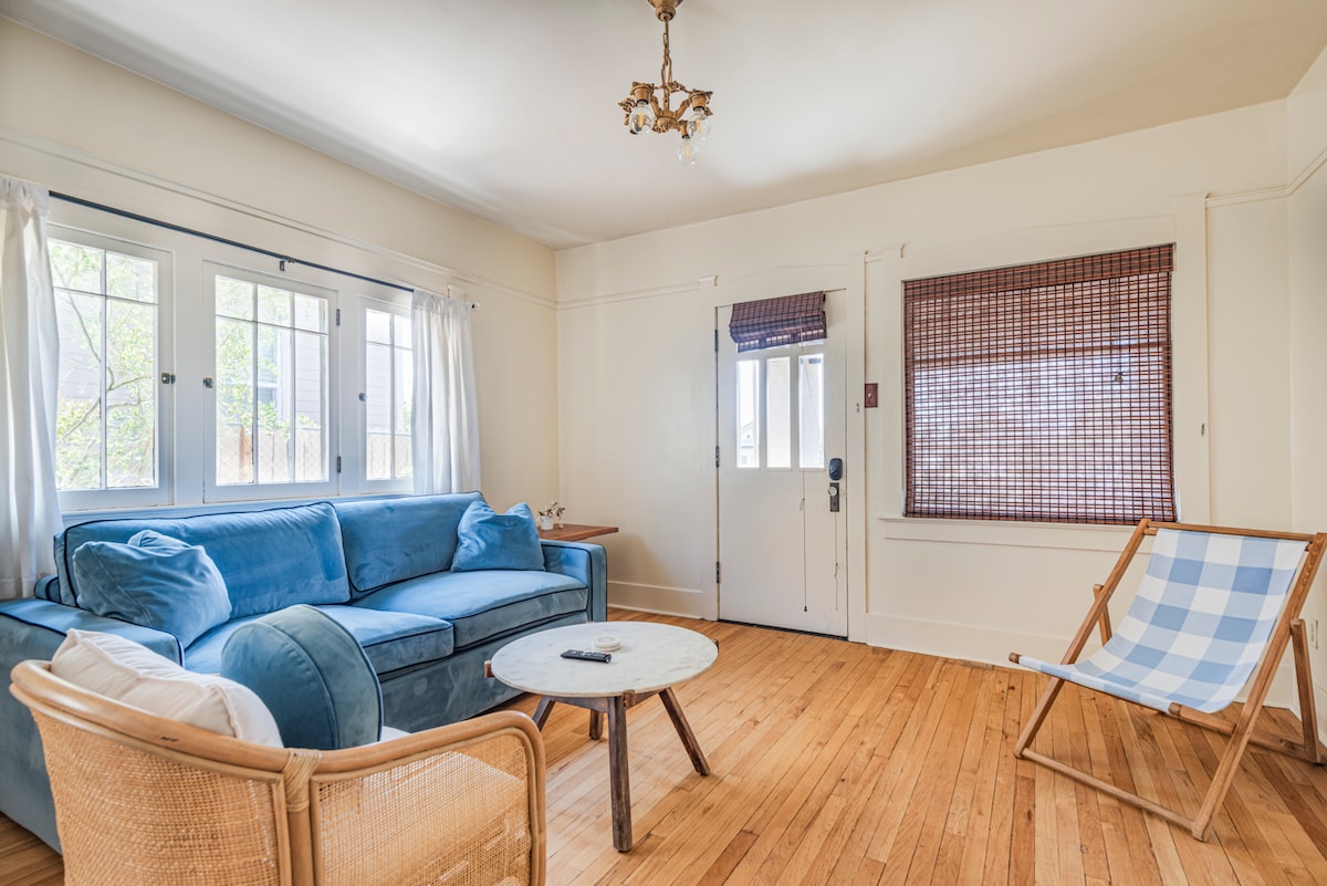 A cozy living room features a blue couch arranged around a round marble table. Natural light filters through large windows, complemented by bamboo shades. A light wood floor adds warmth to the space, which includes a folding chair with a blue and white checkered pattern.