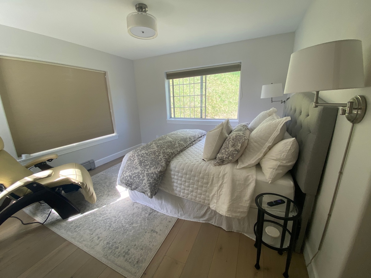 חדר השינה הראשי עם מיטת קווין סייז יוצאת דופן המעוטרת במצעים של אסם קדרות, תאורה צדדית ושולחנות וכיסא מושלם נוסף להירגעות.