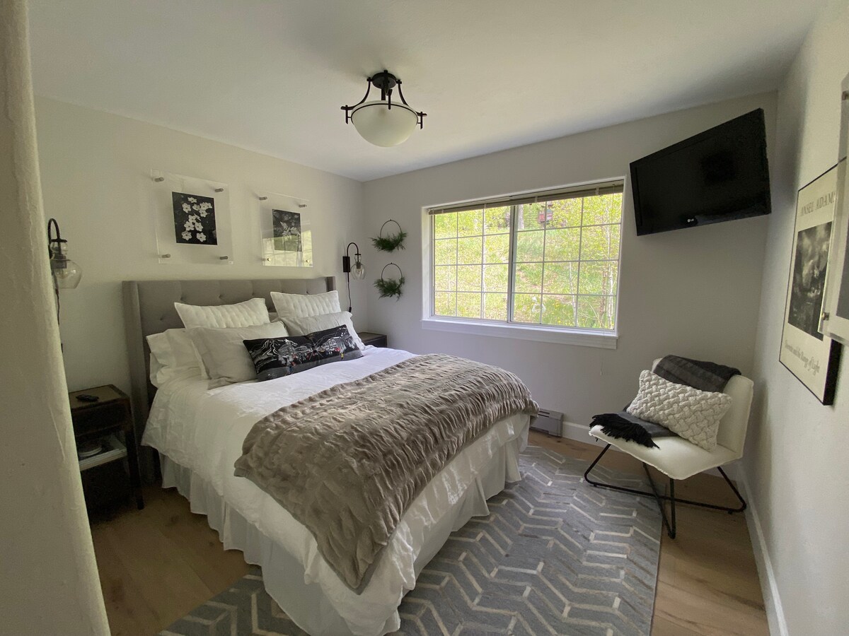 חדר אנסל אדמס. מיטת קווין נוחה עם מצעים, תאורה ושולחנות קצה.  כרית מבטא מדהימה של טלורייד וספרי טליורייד בשולחנות הצדדיים.  אנסל אדמס מצלם נופים אמריקאים איקוניים מקשטים את החדר.