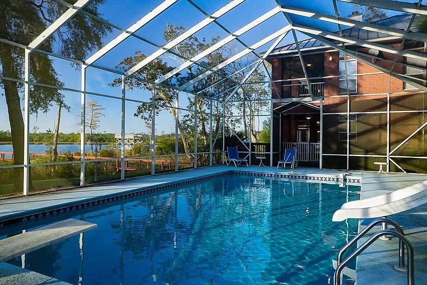 A screened-in pool area features a large swimming pool with crystal clear water. Sun loungers can be seen alongside the pool, and a view of the serene waters of Alaqua Bayou is visible through the enclosure.
