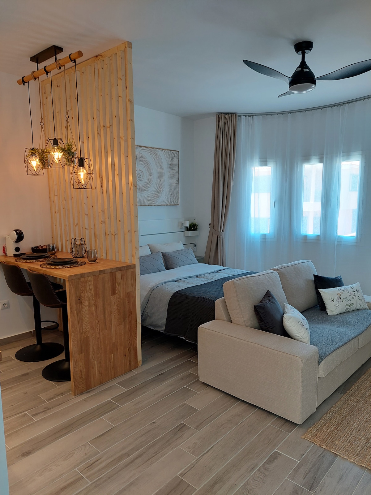 A cozy studio space is featured, with a spacious bed positioned near a set of large windows that allow natural light to fill the room. A small dining area with two stools and pendant lights separates the sleeping area from a comfortable sofa, creating a welcoming ambiance.