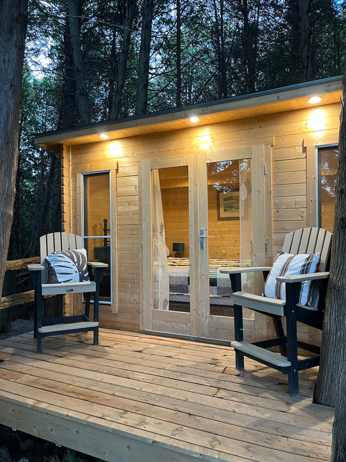 A wooden cabin exterior is surrounded by tall trees, featuring a spacious deck with two black and beige chairs. Large glass doors invite natural light into the interior, where cozy furnishings can be glimpsed. Soft lighting highlights the cabin's welcoming ambiance.