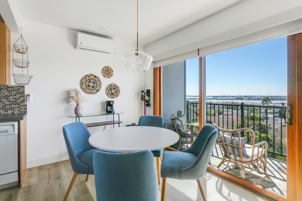 A dining area features a round table surrounded by four blue chairs, with natural light streaming in from sliding glass doors. A small patio with outdoor seating is visible through the doors, offering a view of the bay and city skyline.