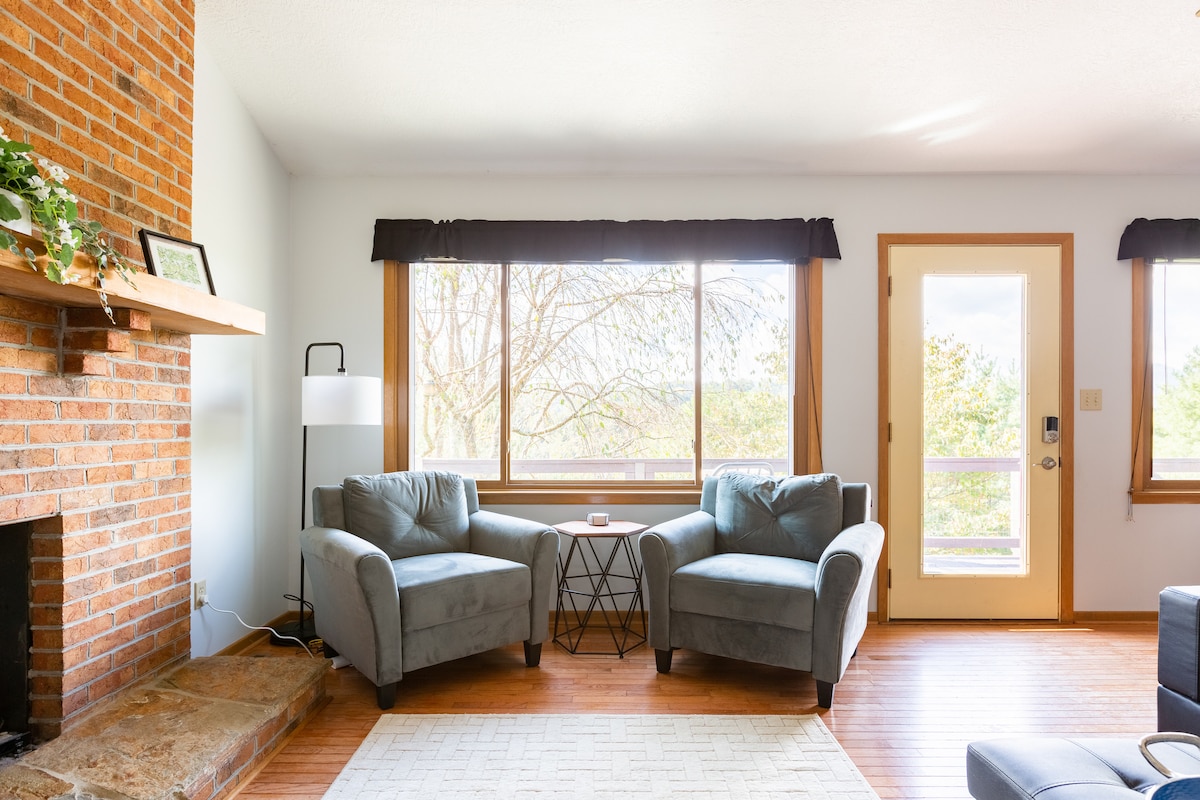 A bright living area features two inviting gray armchairs facing large windows with natural light flowing in. A small table sits between the chairs, and a brick fireplace is positioned against the wall. A door leads outside, enhancing the spacious feel.