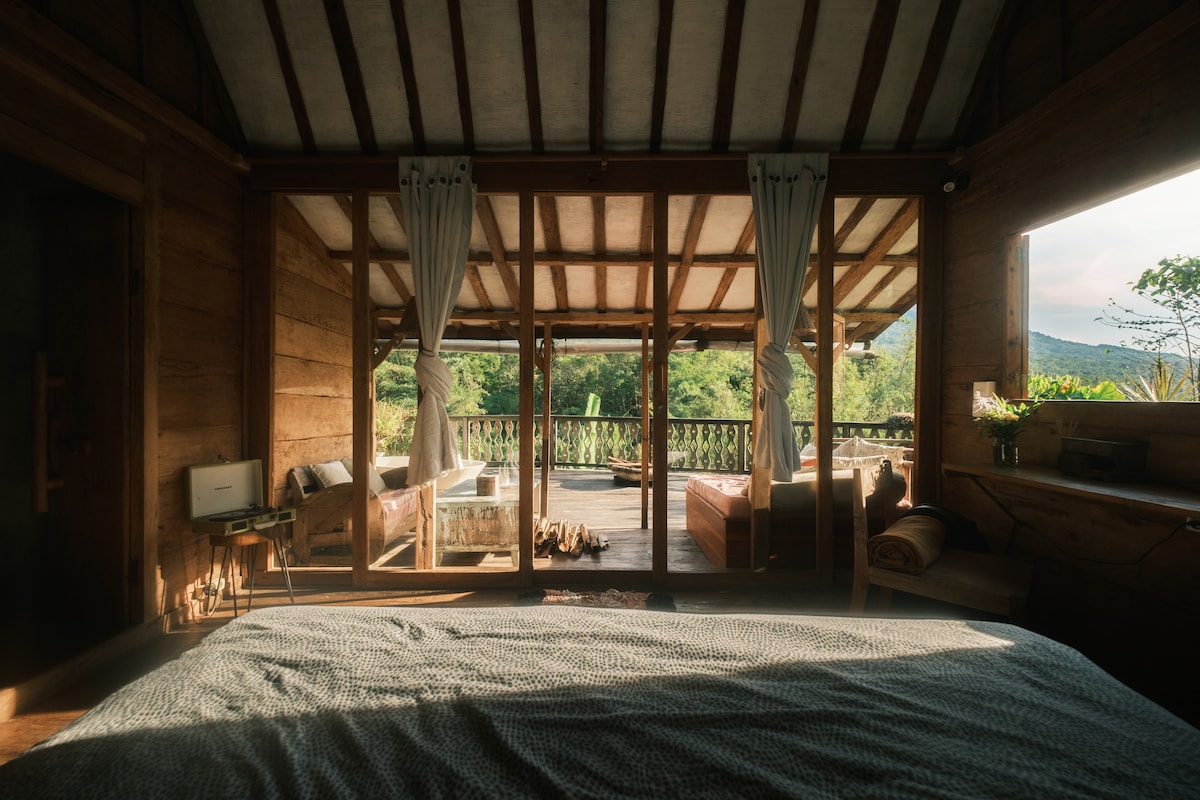 A spacious sleeping area showcases a large bed covered with a soft quilt, inviting restful sleep. Natural light floods the room through expansive glass doors, revealing a terrace overlooking the lush surrounding landscape. Cozy seating areas are visible just outside, creating a welcoming atmosphere.