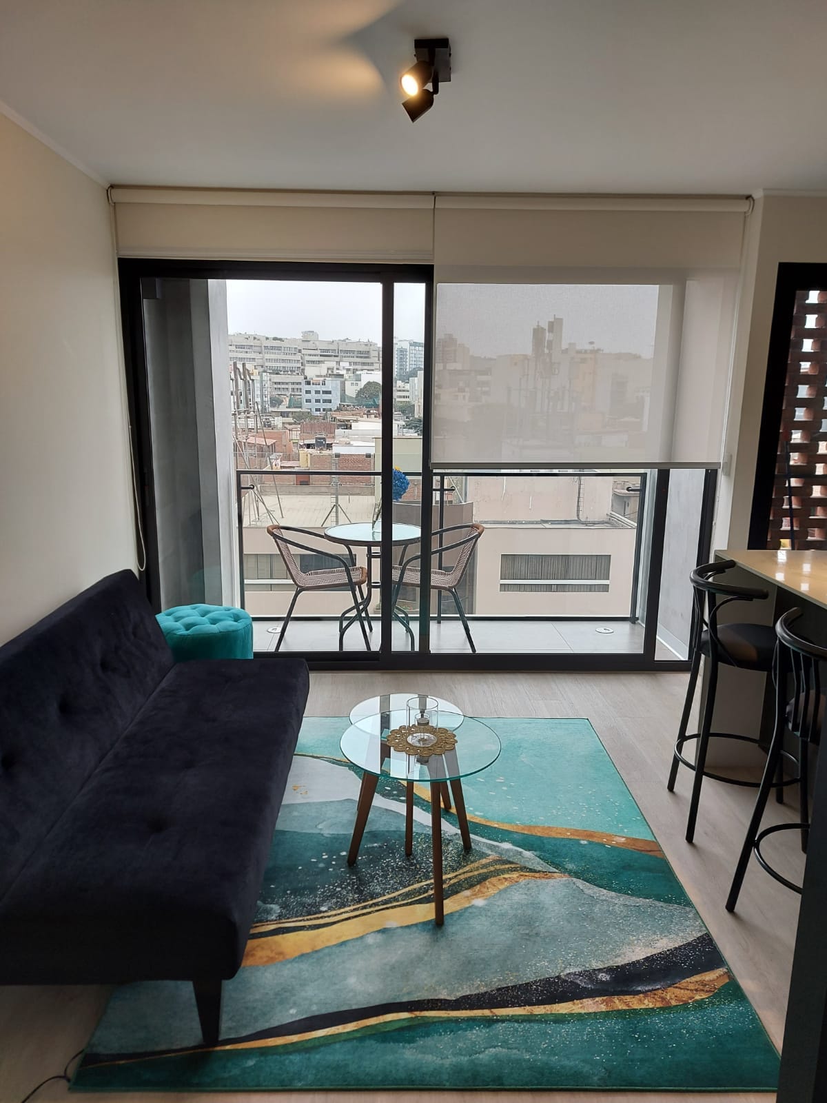 A living area features a dark sofa, a round glass table, and a decorative area rug with abstract patterns. A balcony can be seen, offering views of the surrounding cityscape. Natural light is present through large sliding glass doors.