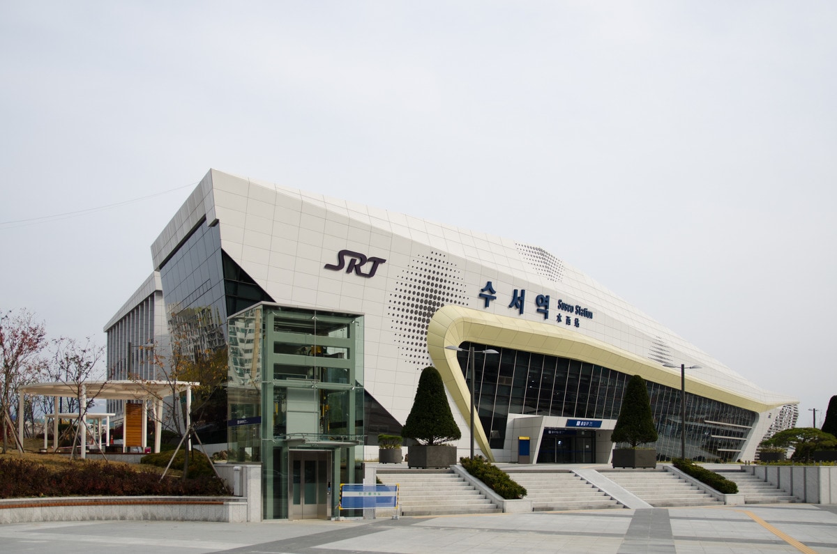 The SRT Suwon Station is depicted with its modern architectural design featuring a sleek, angular facade. Large glass windows create a bright atmosphere, while trees and neatly landscaped areas enhance the exterior. Clear pathways lead to the entrance, inviting easy access for visitors.