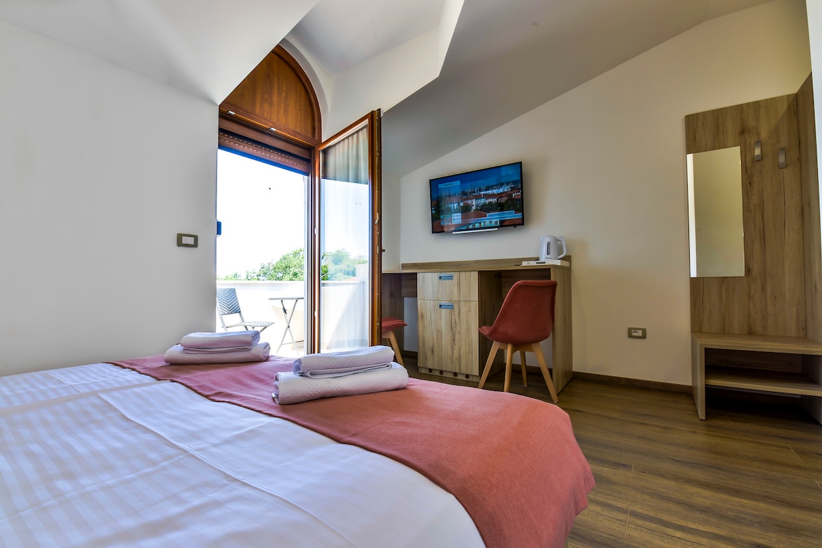 A bright room features two spacious beds covered with soft linens. A modern television is mounted on the wall opposite a small wooden desk and chair. Natural light filters through large windows that open to a balcony with outdoor seating.