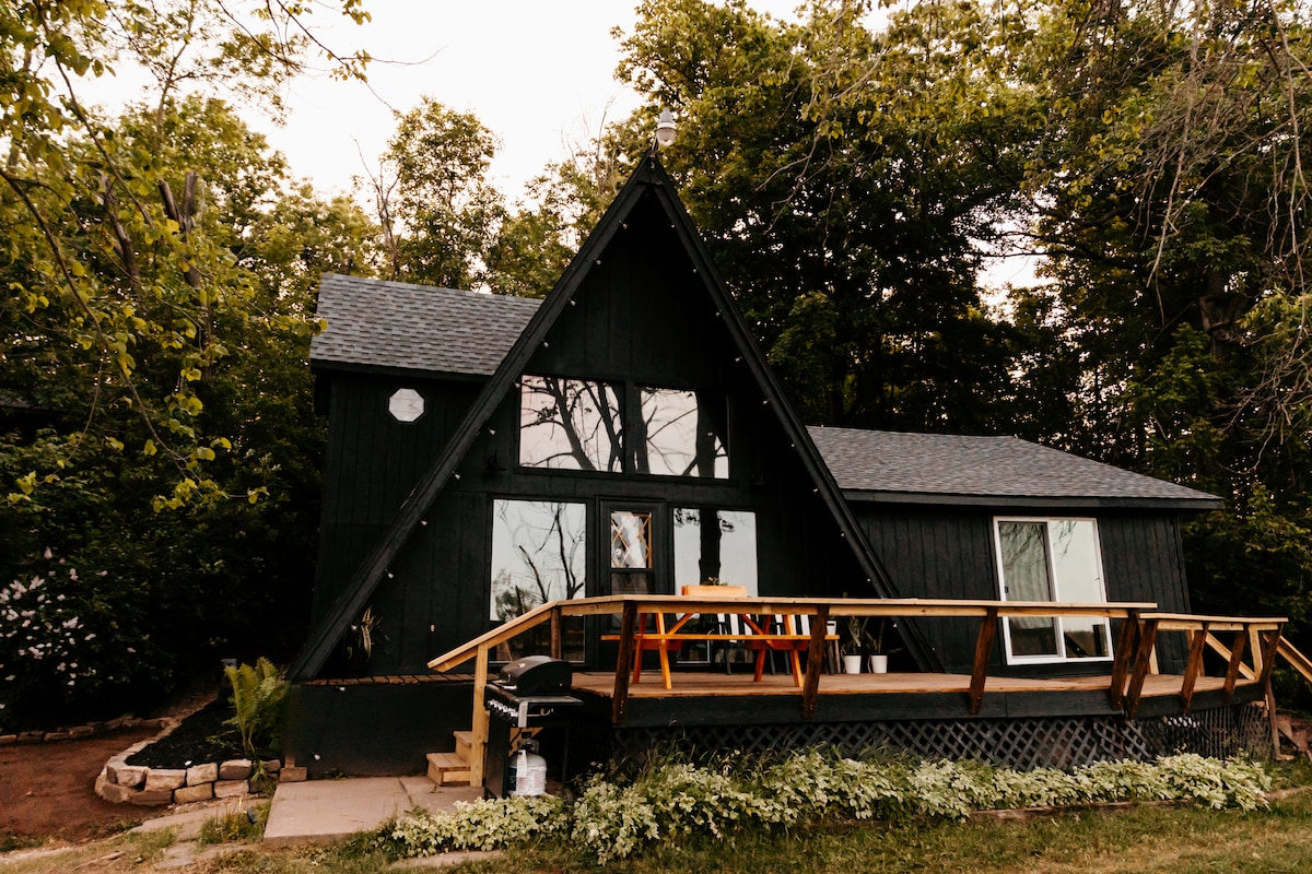 The A-frame cabin is set amidst lush greenery, featuring large windows that invite natural light and offer views of the surrounding landscape. A wooden deck extends from the front entrance, complemented by a seating area and a propane grill, creating an inviting outdoor space.