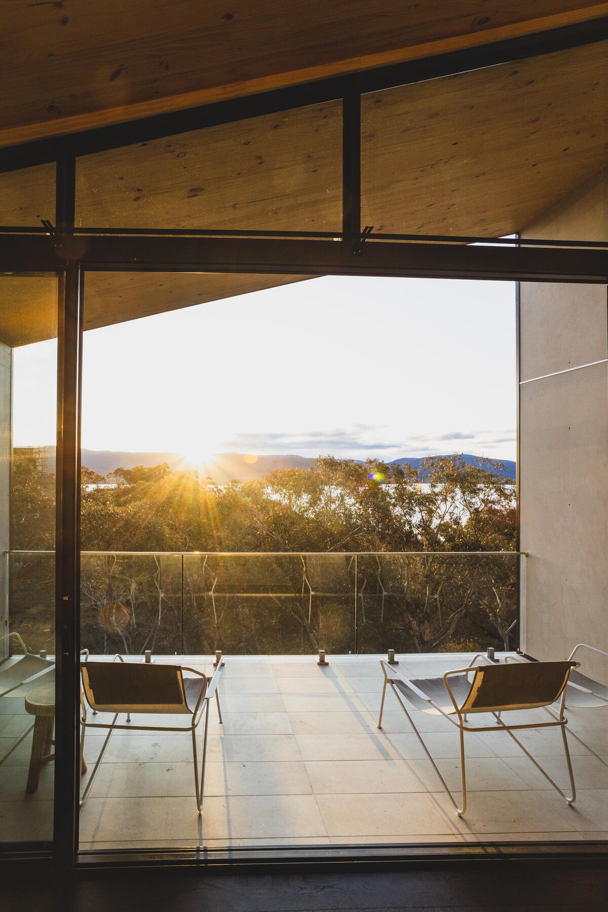 A spacious outdoor balcony is visible, featuring two comfortable chairs facing the sunrise. Large glass doors create a seamless transition between the indoor and outdoor areas, framing an inviting view of the surrounding trees and landscape.