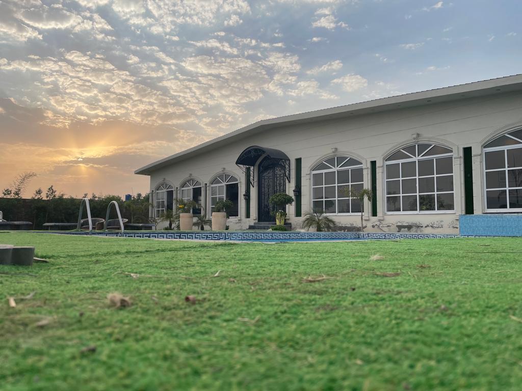 A spacious farmhouse is depicted against a backdrop of a setting sun, with large arched windows framed by green elements. A well-maintained garden and a glistening pool are visible in front, complemented by a grassy area creating a serene outdoor space.