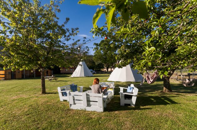 The image captures a tranquil outdoor setting featuring several tipis nestled within a grassy area. A white wooden seating arrangement is visible, surrounded by trees. In the background, hammocks can be seen hanging between the trees, inviting relaxation in the serene natural environment.