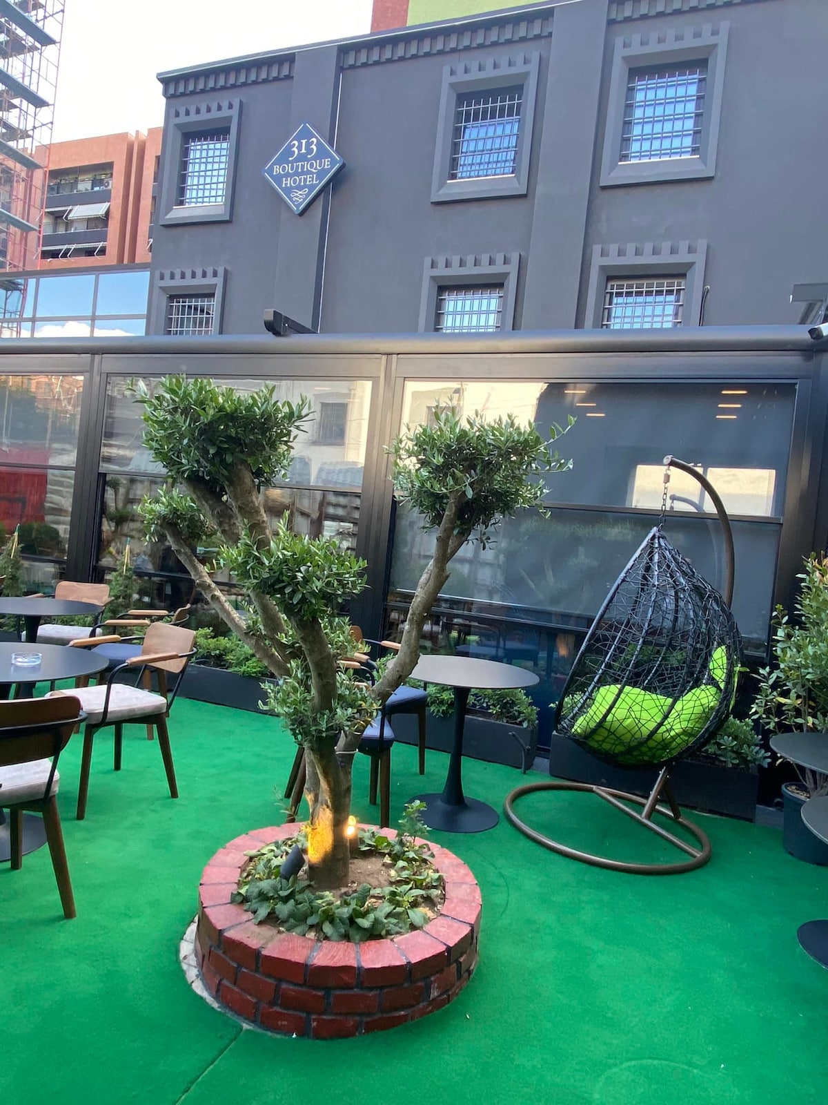 A garden area is shown featuring a vibrant green lawn. Several tables and chairs are placed around an olive tree surrounded by a circular brick border. A hanging wicker chair offers a cozy seating option. The backdrop highlights the modern building's facade.