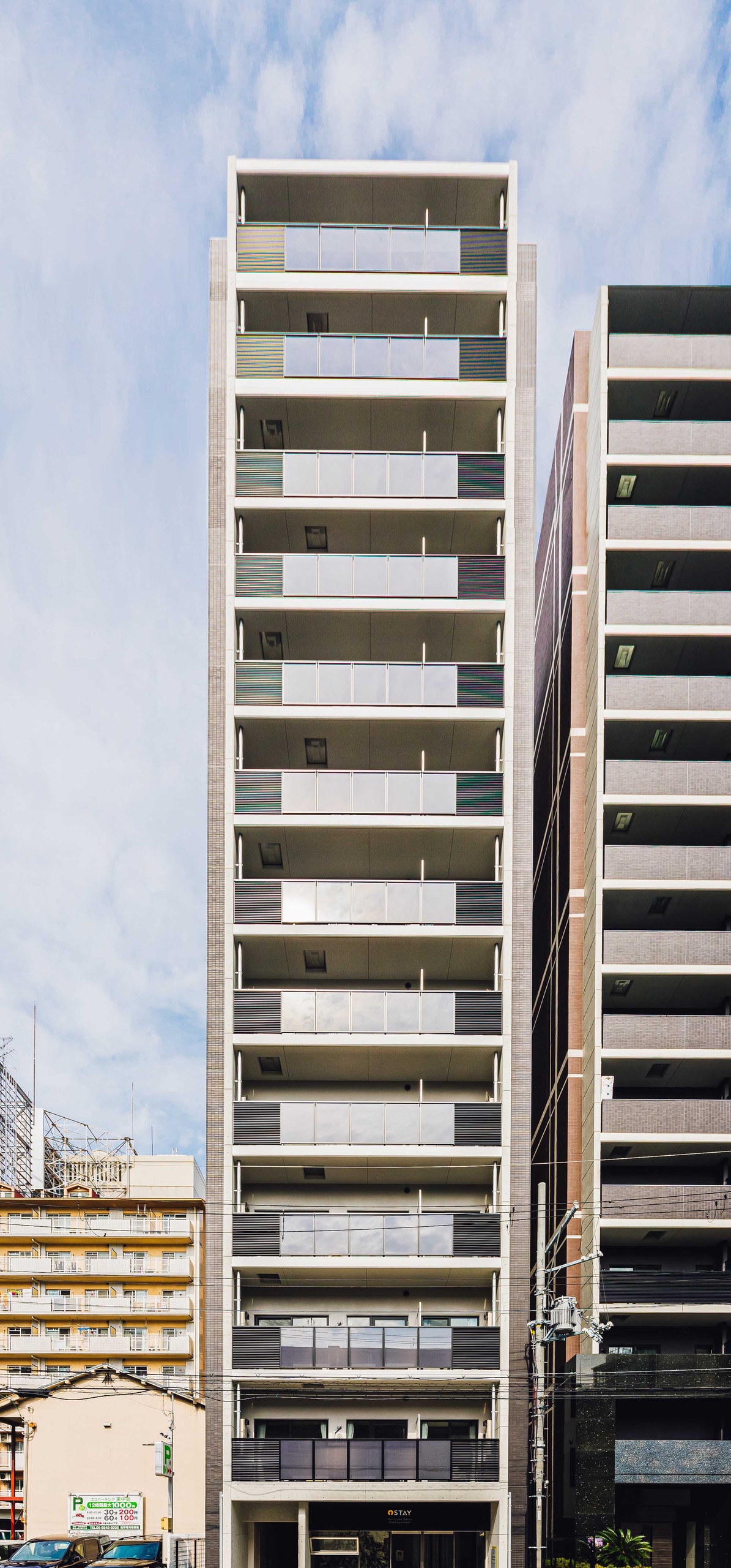 A sleek modern building rises against a blue sky, featuring multiple balconies and large windows on each floor. The façade is a combination of light and dark tones, presenting a contemporary aesthetic. Adjacent buildings offer a glimpse of the urban environment surrounding the property.