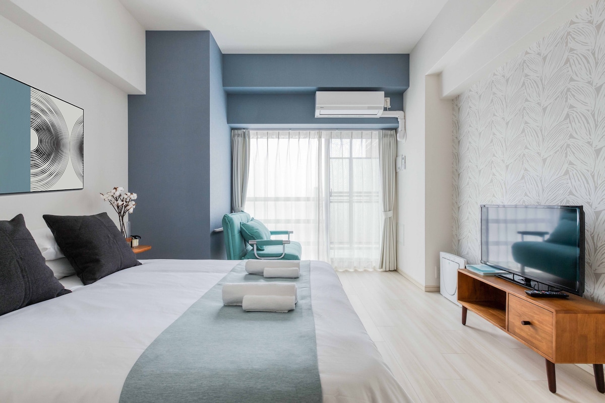 A spacious room features a comfortable double bed with dark pillows and a light-colored duvet. A television is positioned on a wooden console, and a chair in the corner complements the blue accent wall. Natural light filters through sheer curtains, enhancing the airy feel of the space.