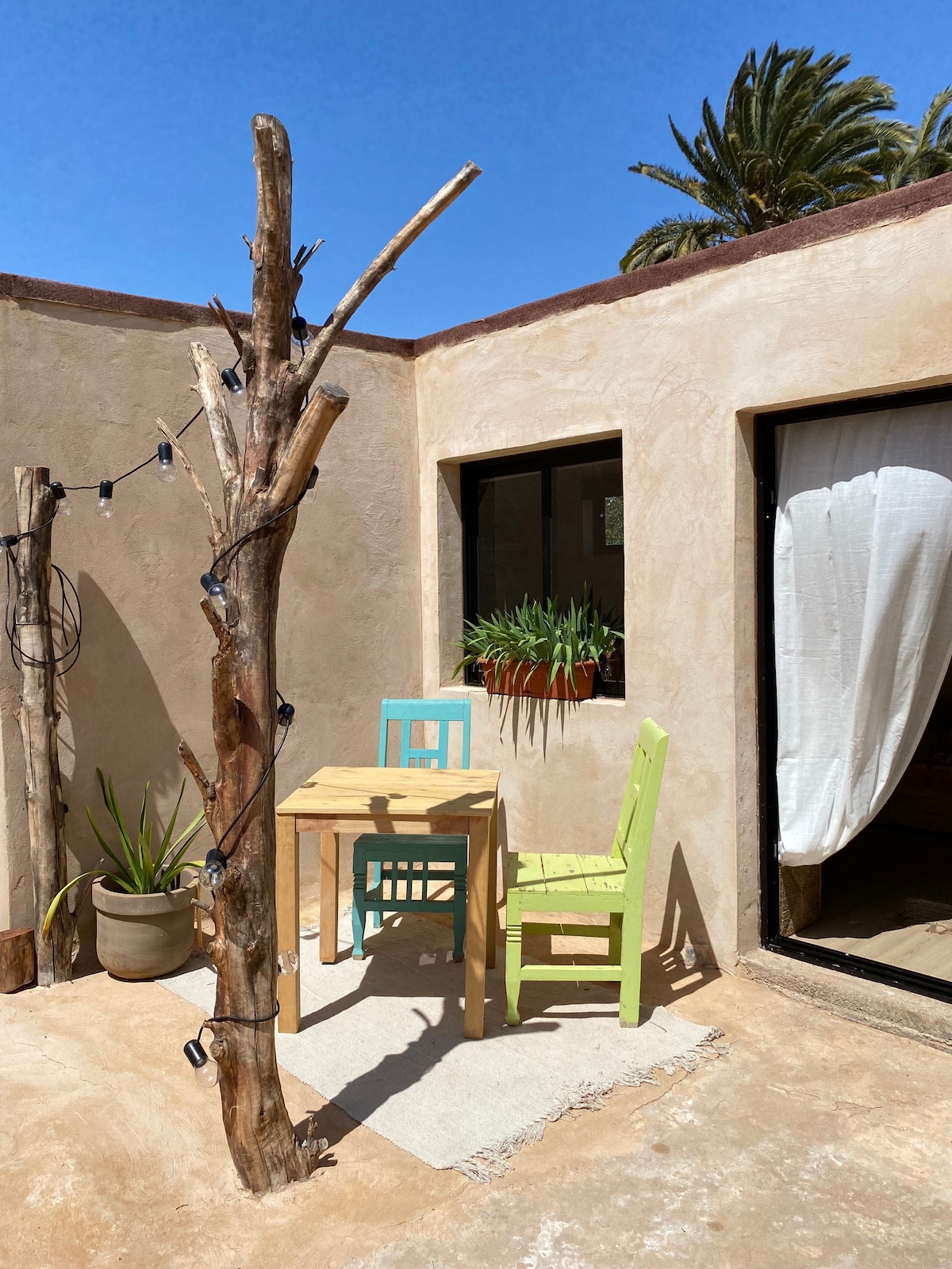 A quaint outdoor area is presented, featuring a small wooden table surrounded by two colorful chairs in green and blue. Natural light fills the space, with potted plants adding greenery. A tall, rustic tree trunk is visible, enhancing the serene ambiance.