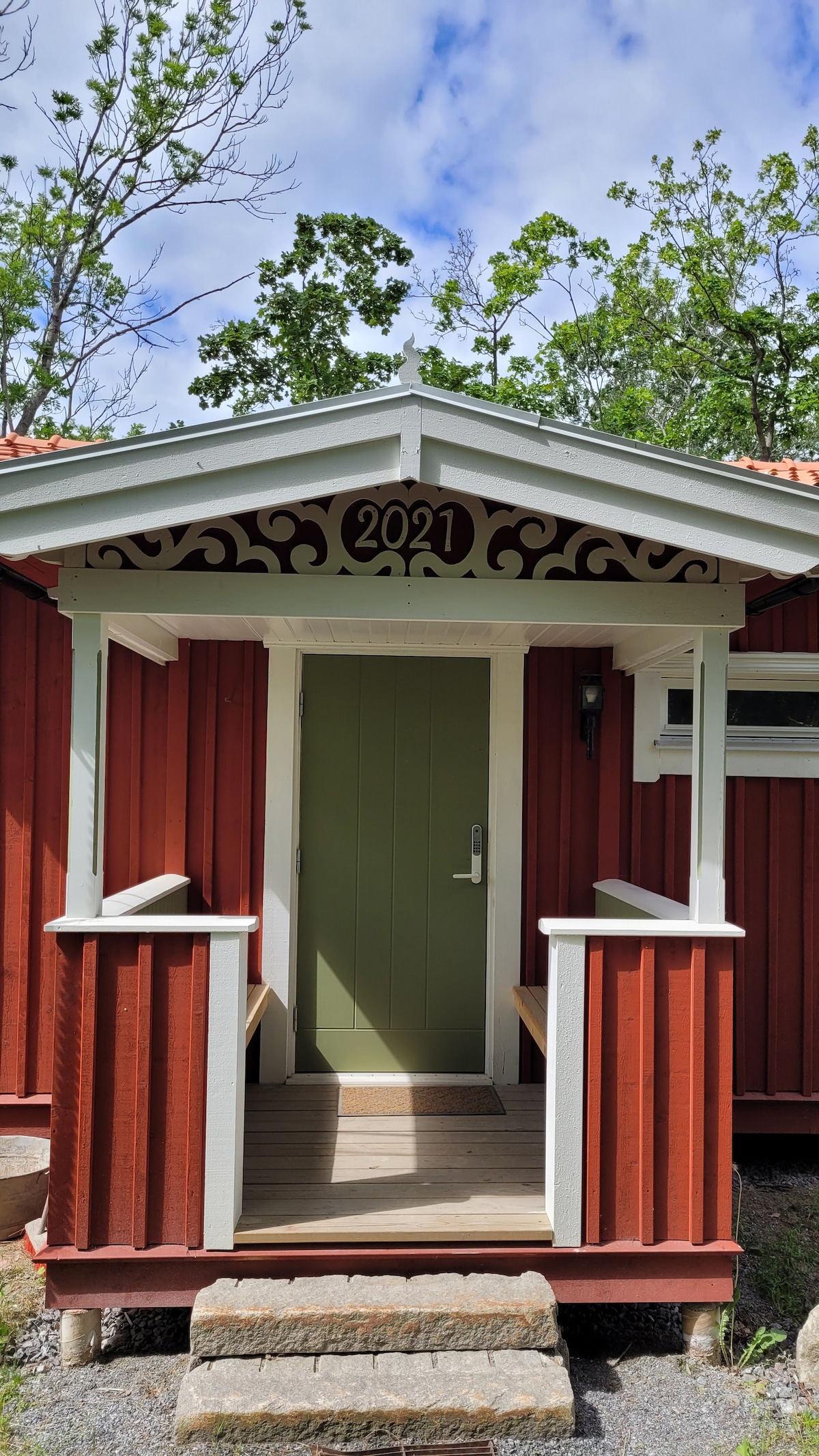 The entrance to the house showcases a red wooden facade with white trim and a charming roof overhang. A green door is framed by two benches on either side, offering a welcoming place to sit. The year '2021' is prominently displayed above the door.