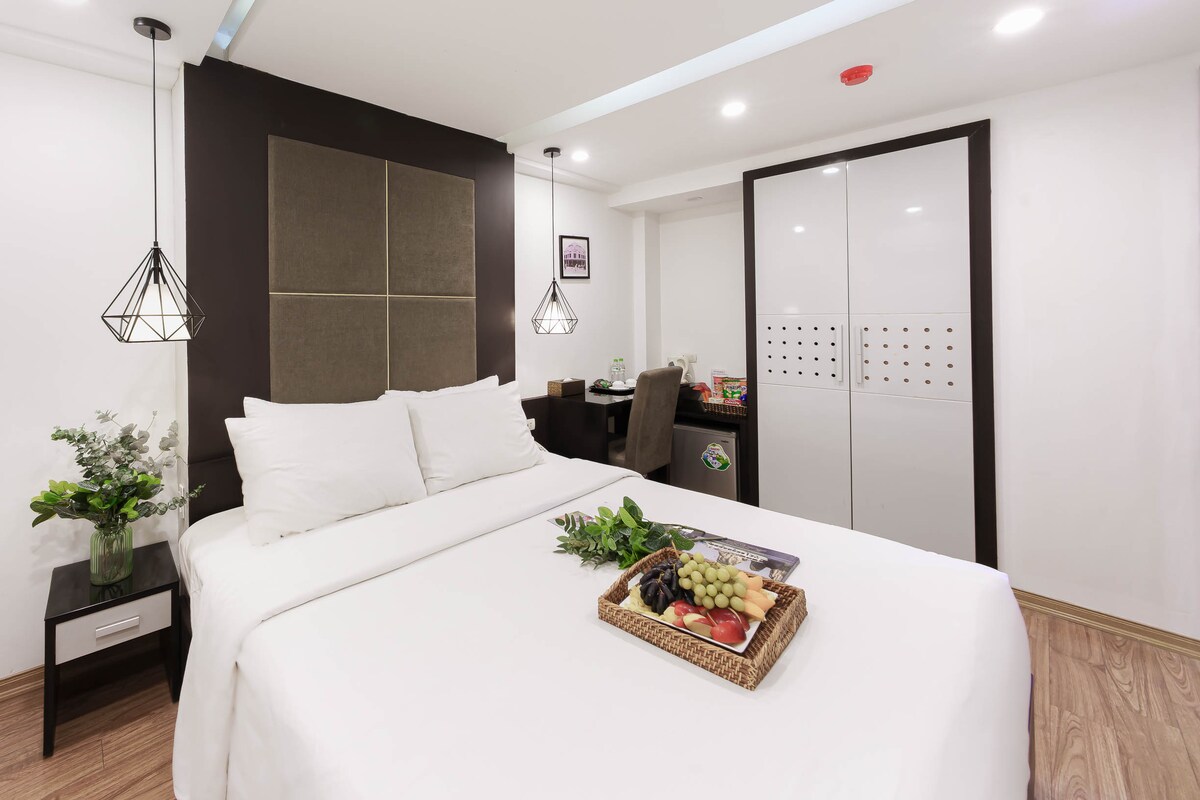 A comfortable room features a spacious bed adorned with crisp white linens. A small basket of fresh fruit is placed on the bed, while a sleek desk and storage area are visible in the background. Soft lighting is provided by stylish pendant lamps.