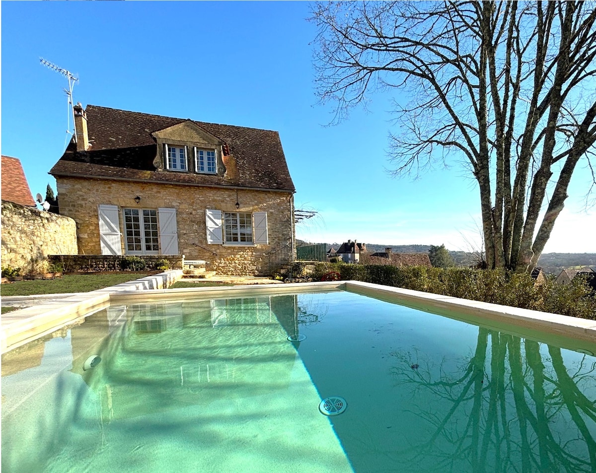 A sunlit view of the charming stone house is captured from the edge of the swimming pool. Large windows reflect a serene landscape, while a blooming tree stands nearby. The pool's clear water mirrors the blue sky, emphasizing the peaceful setting.